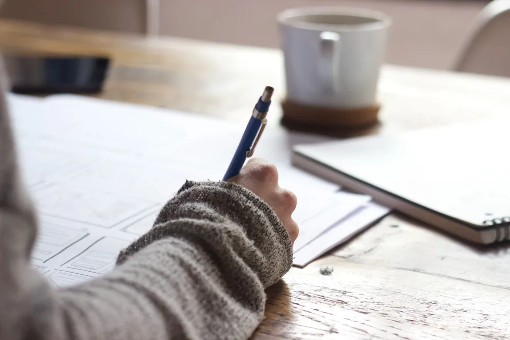 Hand holding a blue pen over papers on a table beside a mug