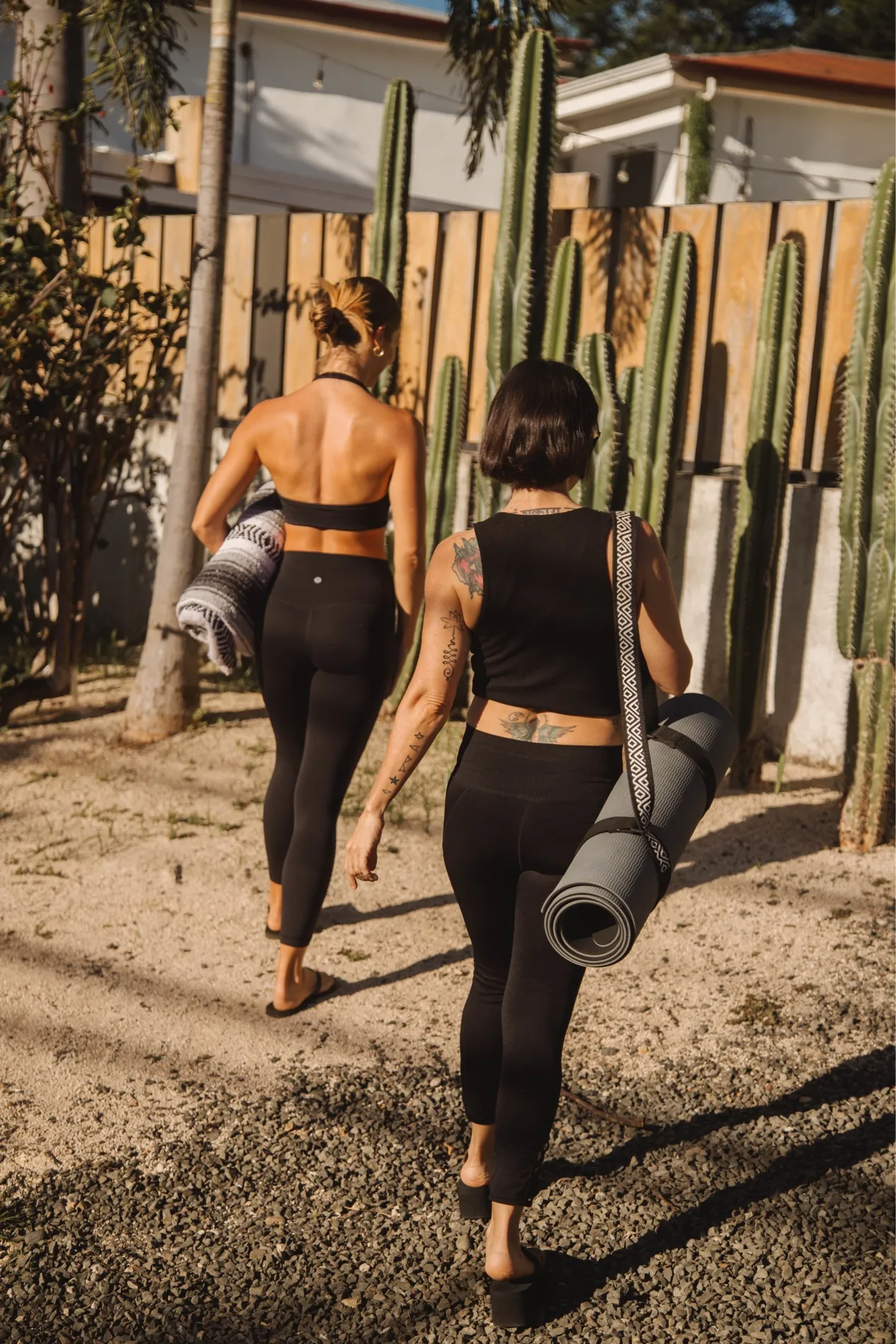 Two women in black workout wear walk past a cactus fence carrying rolled yoga mats