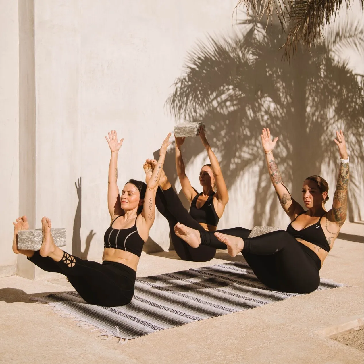 Three women in black sit balanced in a Pilates pose with arms raised beside a palm shadow on a wall