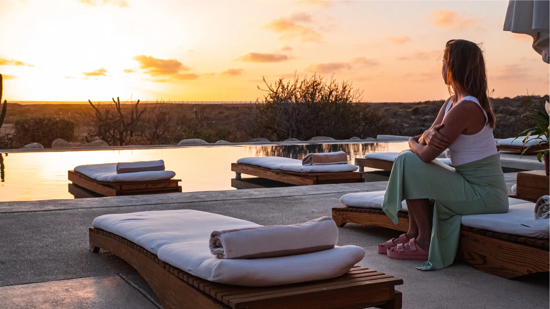 Woman in a green skirt sits on a lounger by a pool at sunset
